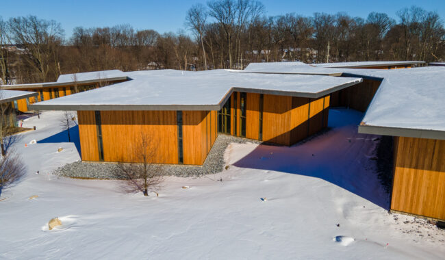 Modern wooden buildings with angular roofs sit among snow-covered ground and leafless trees under a clear blue sky. The sunlight casts shadows, highlighting the winter landscape and the unique architecture.