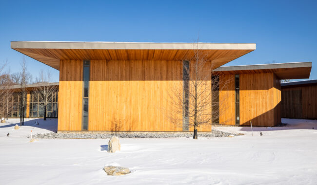 Modern wooden building with tall, narrow windows and a flat roof, surrounded by snow. A small leafless tree and rocks are in the snowy foreground under a clear blue sky.