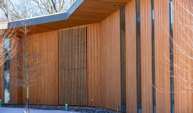 A modern building exterior with vertical wooden paneling, tall narrow windows, and a snow-covered ground. Leafless trees stand near the building, and rocks line the base of the structure.