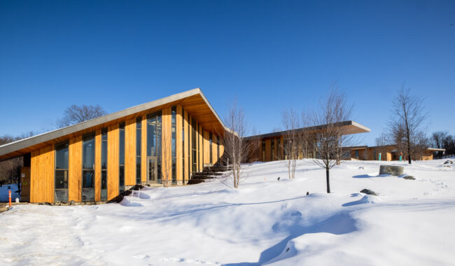 A modern wooden building with large glass windows and sloped roofs sits amid a snowy landscape with bare trees under a clear blue sky.