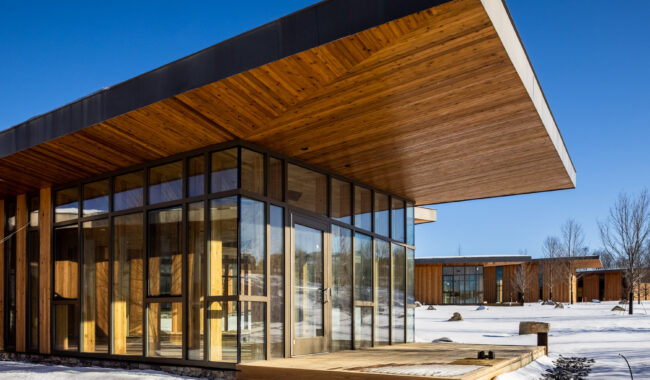 A modern building with large glass walls and an expansive wooden overhang stands on a snowy landscape under a clear blue sky. Another similar structure is visible in the background among bare trees.