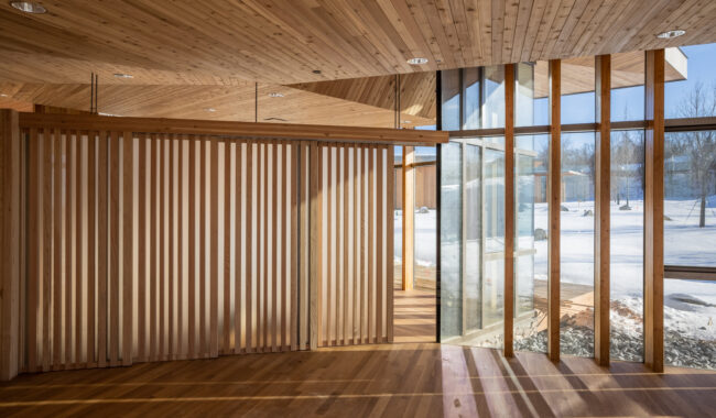Sunlight streams into a modern room with wooden floors, walls, and ceiling. Vertical wooden slats and tall glass windows reveal a snowy outdoor landscape and trees. The space feels warm and open.