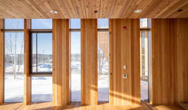 Sunlight streams through tall, narrow windows in a modern room with wooden floors, walls, and ceiling. Outside, snow covers the ground, and bare trees stand against a bright winter sky.