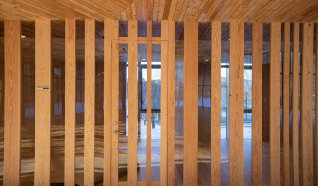 A wooden wall with evenly spaced vertical slats and a glass door in the center, revealing a sunlight-filled room with large windows and a snowy landscape outside.