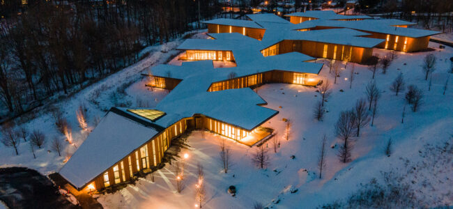 Aerial view of a modern building with angular, branching design, illuminated from within at dusk, surrounded by snow-covered ground and bare trees.