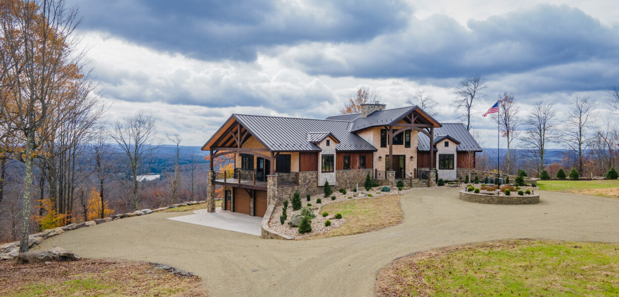Large modern house with stone and wood exterior, set on a hilltop with a winding gravel driveway, surrounded by autumn trees and an American flag, under a cloudy sky.