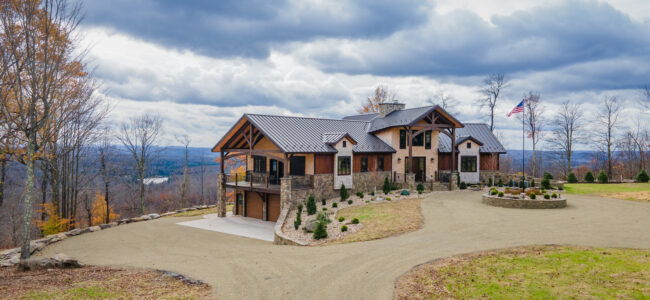 Large modern house with stone and wood exterior, set on a hilltop with a winding gravel driveway, surrounded by autumn trees and an American flag, under a cloudy sky.