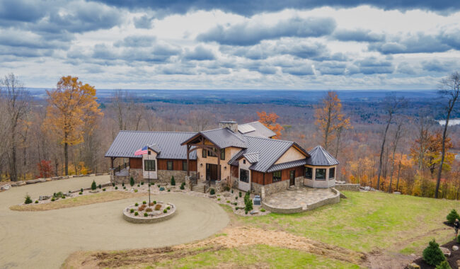 A modern house with stone and wood exterior sits atop a hill, surrounded by autumn trees. A circular driveway with an American flag leads to the entrance. Vast forested landscape and cloudy sky stretch out in the background.