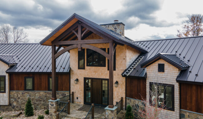 A modern house with wood siding, stone accents, a metal roof, and a large covered entryway supported by timber beams; cloudy sky in the background.