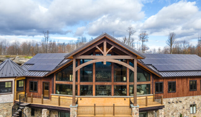 A large modern house with a stone and wood exterior features a prominent glass facade, metal roof with solar panels, and a spacious balcony, set against a backdrop of leafless trees and a cloudy sky.