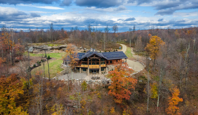 A large log cabin with solar panels on the roof sits among autumn trees and a rocky hillside under a cloudy sky, with a winding gravel driveway leading to the house.