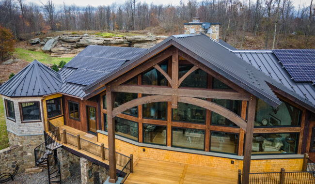 A modern house with large glass windows, wooden beams, a spacious deck, and rooftop solar panels, set in a wooded, rocky landscape with trees and overcast skies in the background.