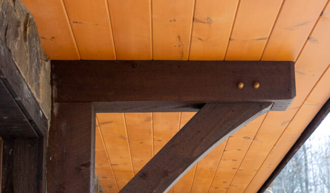 A close-up view of a wooden support bracket attached to a stone wall, with a light orange, wood-paneled ceiling above and trees visible outside in the background.