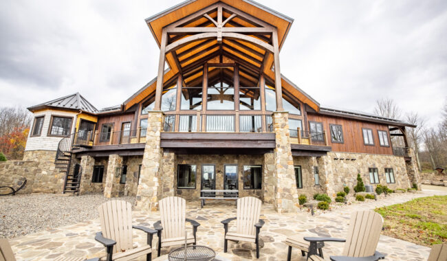 A large stone and wood lodge with a high, peaked roof, large windows, and a patio with Adirondack chairs arranged around a fire pit, surrounded by trees under a cloudy sky.