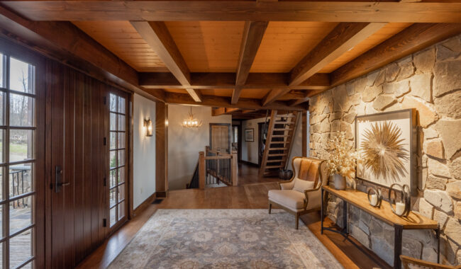 A cozy entryway with exposed wooden beams, stone accent wall, two armchairs, a rug, and decorative art. Glass doors lead outside, and sunlight brightens the space with a warm, inviting atmosphere.
