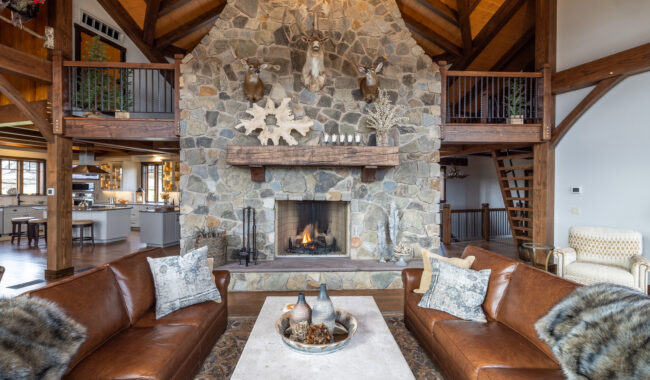 A rustic living room with a tall stone fireplace, mounted deer heads, wooden beams, brown leather sofas, and a modern kitchen visible in the background. A fire is burning in the fireplace.