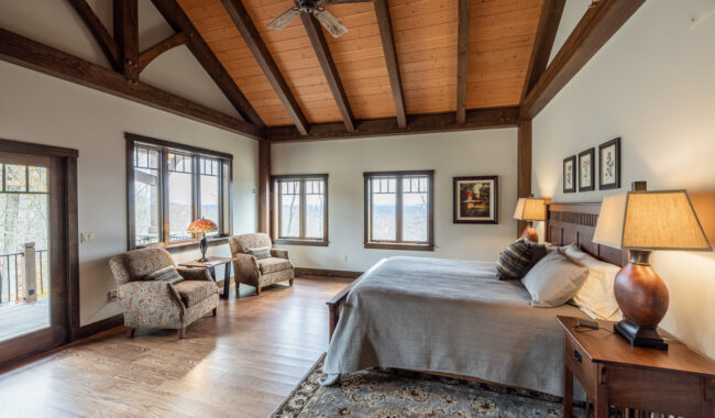 Spacious bedroom with a vaulted wood beam ceiling, large windows, a king bed with gray bedding, two patterned armchairs by the window, lamps, framed art, and a rug on hardwood floors. Balcony door visible on the left.