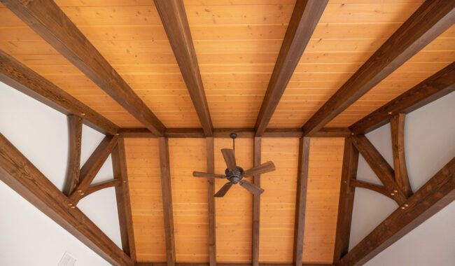 Ceiling view of a room with exposed dark wooden beams, light wood paneling between the beams, and a ceiling fan with five blades in the center. The room has white walls and recessed lighting fixtures.