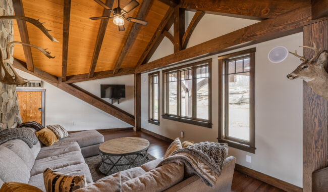 A cozy living room with a high wooden ceiling, large windows, a sectional sofa with pillows and throws, a round coffee table, a mounted TV, and deer mounts on the walls. Warm natural light fills the space.