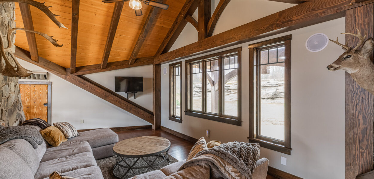 A cozy living room with vaulted wooden ceilings, exposed beams, deer head mounts, large windows, a sectional sofa with pillows and blankets, a round coffee table, and a wall-mounted TV.