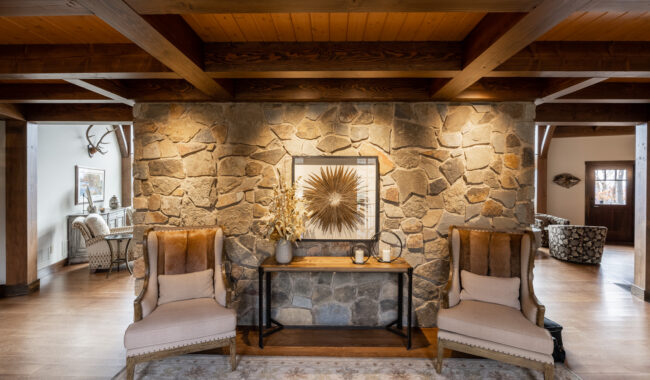 A cozy living space with a stone accent wall, two tan armchairs with wooden sides, a narrow table with decorative items, sunflower art, wooden beams on the ceiling, and warm lighting.