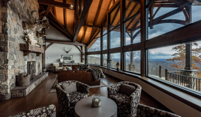 A rustic living room with high wooden ceilings, large floor-to-ceiling windows overlooking a scenic mountain view, stone fireplace, animal mounts, cozy seating, and a patterned armchair set around a round table.