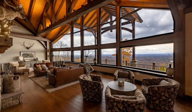 Spacious living room with large floor-to-ceiling windows offering a scenic view of mountains and forest. The room features wooden beams, leather sofas, patterned armchairs, and rustic decor with warm lighting.