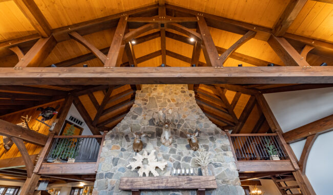 A rustic living room with a high vaulted wooden ceiling, exposed beams, a large stone fireplace, mounted deer heads, and decorative accents on the mantel. Balconies are visible on either side above the fireplace.