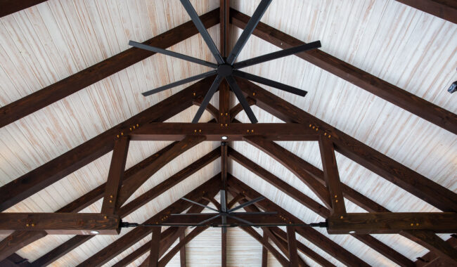 A large black ceiling fan with ten blades is mounted on a vaulted ceiling featuring dark wooden beams and light-colored wooden planks.