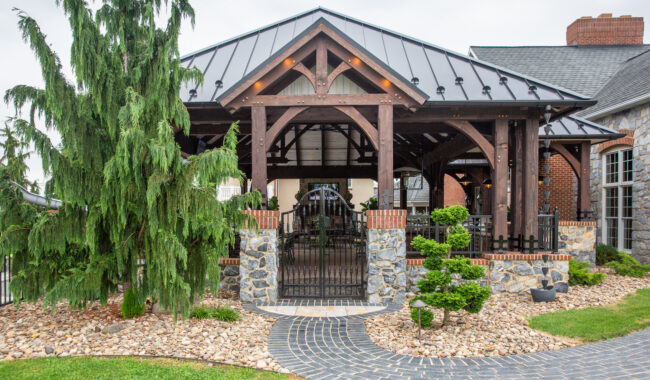 A wooden pavilion with a metal roof and stone pillars, surrounded by landscaped rocks, shrubs, and a large evergreen tree, with a curved brick pathway leading to the entrance.