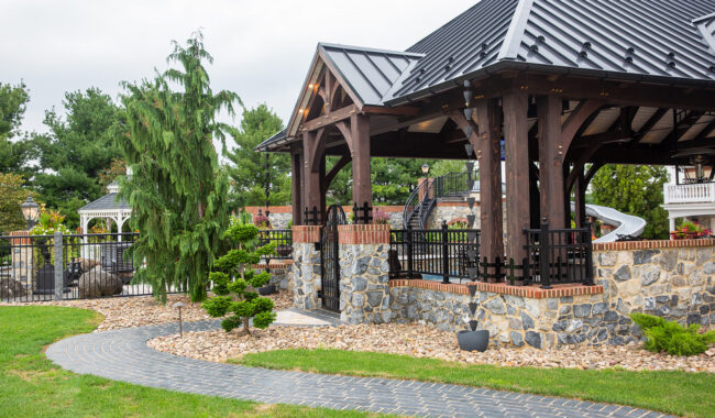 A stone pathway curves through a landscaped yard toward a wooden pavilion with a black metal roof, surrounded by greenery and trees under a cloudy sky.