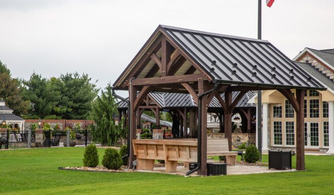 A wooden pavilion with a metal roof stands on a green lawn near a building. Benches are arranged underneath the structure, and a U.S. flag is visible on a pole in the background. Shrubs and trees border the area.