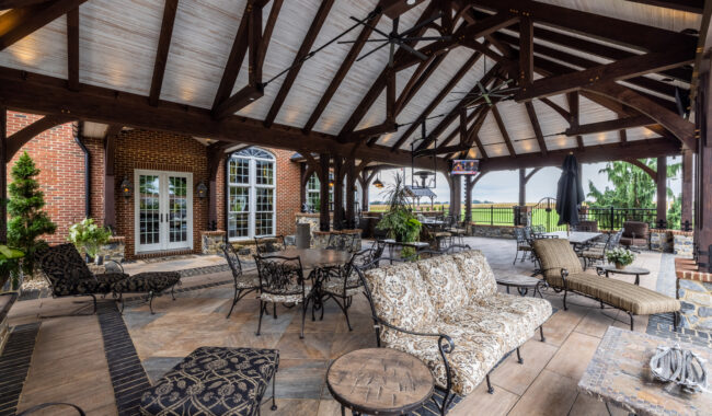 Elegant covered patio with exposed wooden beams, cushioned seating, metal tables and chairs, potted plants, and a view of the outdoors; brick wall and large windows in the background.