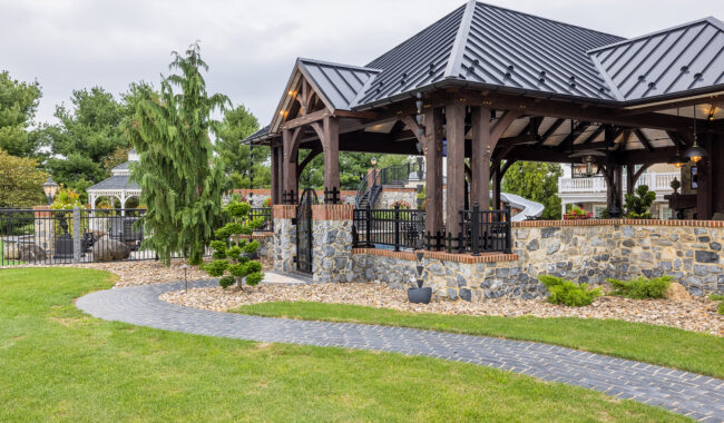 A large outdoor pavilion with a black metal roof, stone and brick pillars, and black fencing is surrounded by landscaped grass, a curving brick pathway, and small trees on a cloudy day.