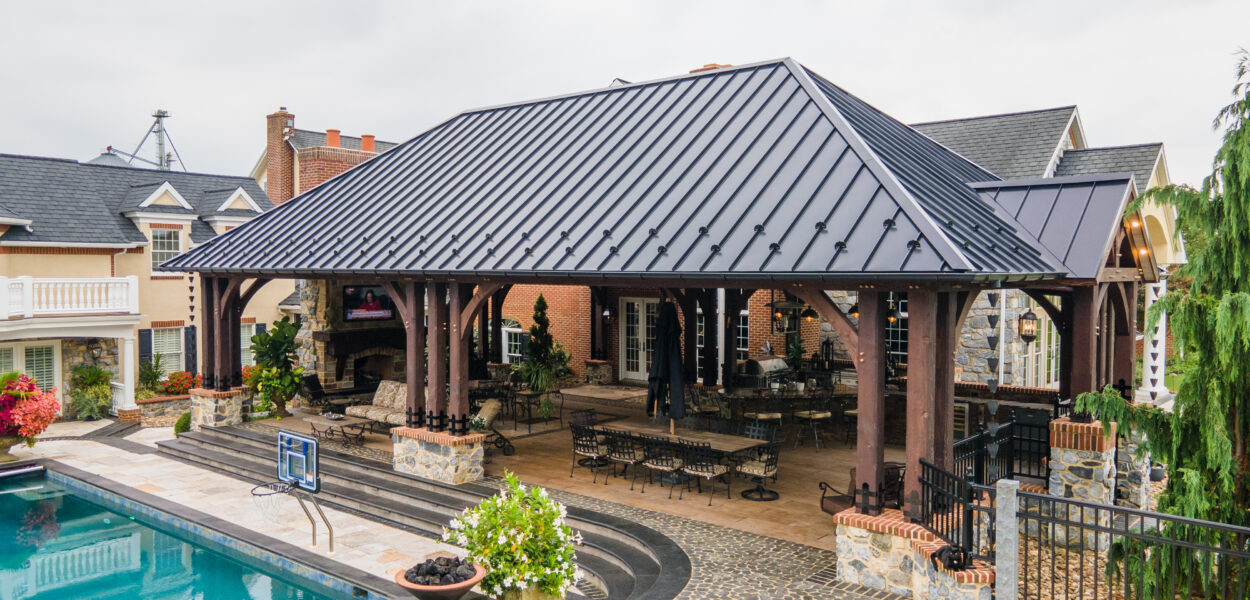 A large covered patio with a black metal roof and wooden beams stands next to a swimming pool, surrounded by stone paving, outdoor seating, and landscaped greenery at a spacious residential property.
