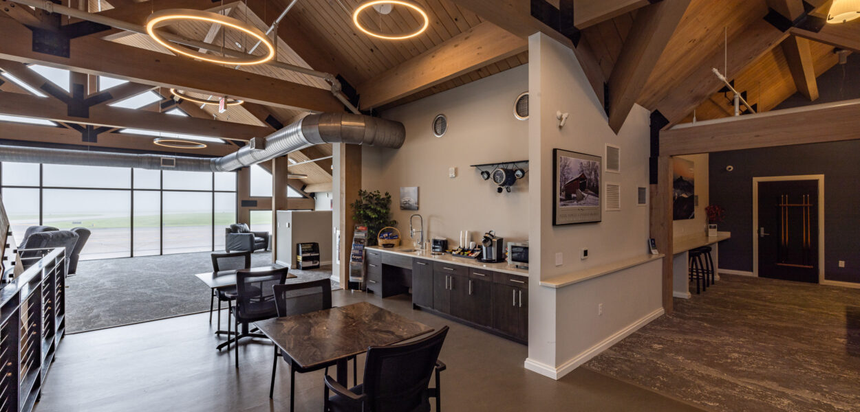 A modern lounge area with high wooden ceilings, circular pendant lights, tables and chairs, a coffee station, and large windows providing natural light. The space has a contemporary, open design.