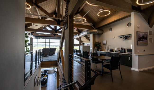 Modern loft-style office space with exposed wooden beams, large windows, a kitchenette, tables, chairs, and industrial lighting, featuring a view of greenery outside.