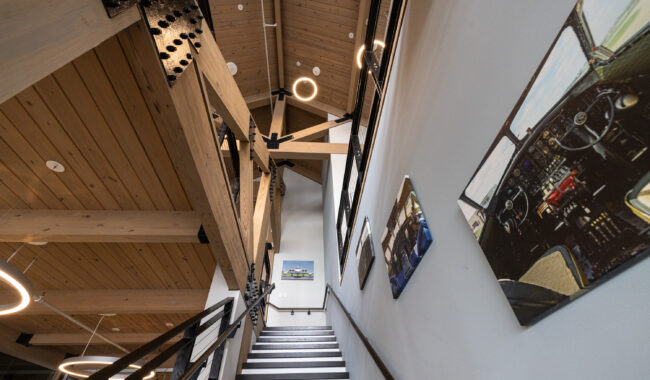 A modern staircase with black and white steps leads upward, flanked by wooden beams and a high, slanted ceiling. Circular lights hang above, and aviation-themed photos are displayed on the wall beside the stairs.