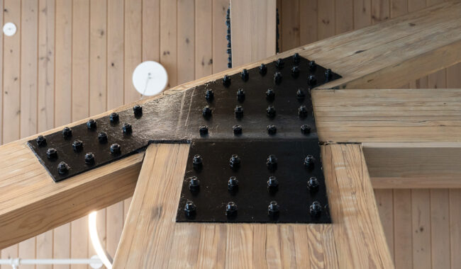 Close-up of wooden beams joined with large black metal plates and numerous bolts, set against a wooden ceiling with visible planks and a circular white light fixture in the background.