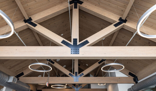 Exposed wooden beams with metal connectors form a vaulted ceiling in a modern interior. Circular pendant lights hang down, and metal air ducts are visible along one side. The space has a clean, contemporary look.