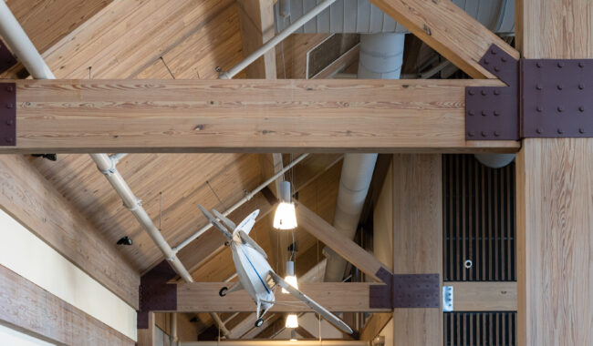 A model airplane is suspended from the wooden beams of a high ceiling with exposed trusses and hanging lights in an indoor space.