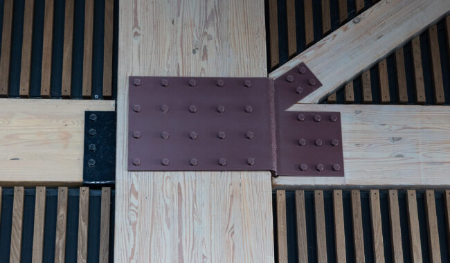 Close-up of a wooden beam structure joined by a large brown metal plate with multiple bolts; dark vertical slats form the background.