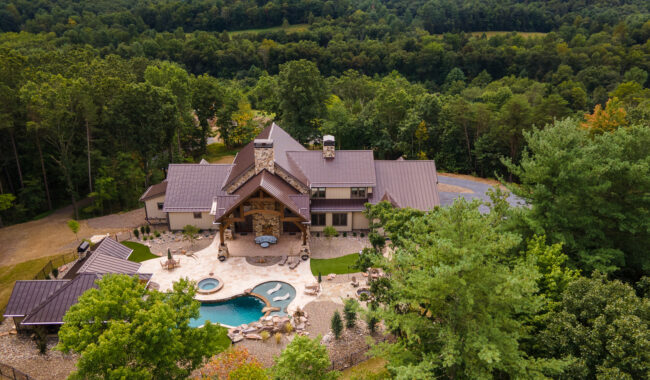 Aerial view of a large, modern house with a metal roof surrounded by dense green trees, featuring a spacious stone patio, swimming pool, hot tub, and landscaped garden.