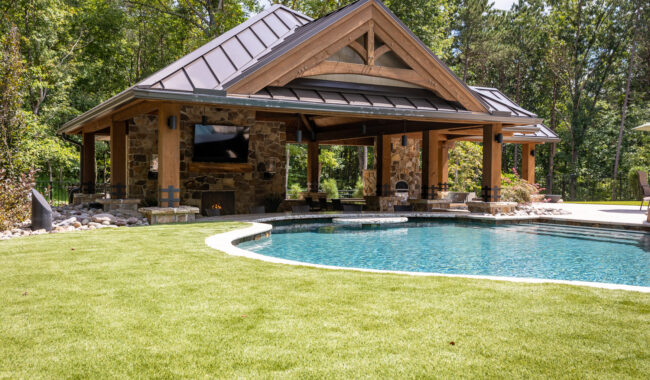 A modern outdoor pavilion with a stone fireplace and mounted TV sits beside a curved swimming pool, surrounded by green artificial grass and trees in a backyard setting.