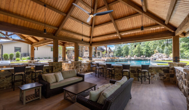 Spacious outdoor pavilion with wooden beams, comfortable wicker seating, tables, bar stools, and a view of a swimming pool surrounded by trees and stone structures under a clear sky.
