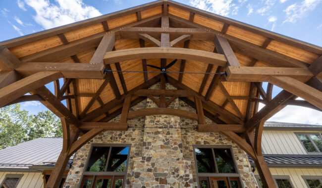 A close-up view looking up at a houses large wooden gabled roof with exposed beams, supported by a stone wall with tall windows below, against a blue sky with clouds.
