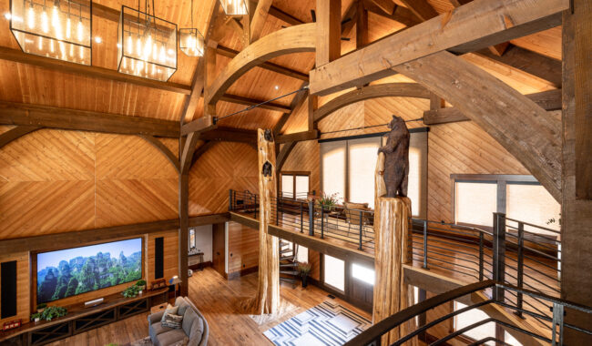 A spacious, rustic living room with exposed wooden beams, high ceilings, a large TV, and a balcony. A carved bear statue sits atop a log pillar, and natural light streams in through large windows.