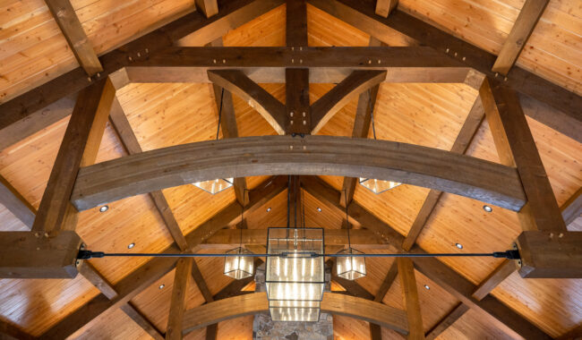 Wooden ceiling with exposed beams and trusses, featuring a central rectangular chandelier and warm lighting; the ceiling panels are light-colored wood, and there is a stone wall partially visible at the bottom.
