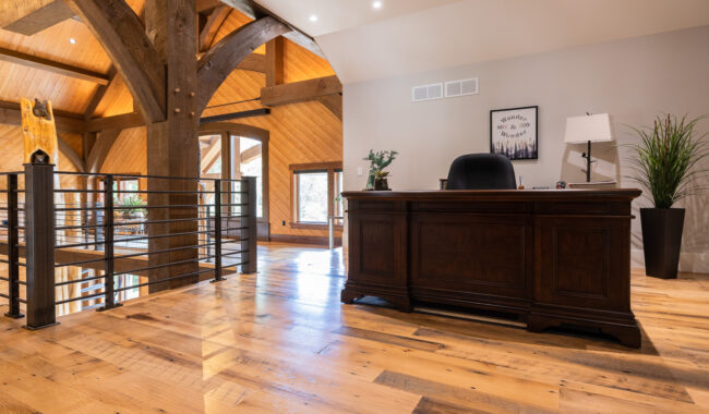 A spacious, modern office with wooden beams, polished wood floors, a large dark wooden desk, potted plants, and natural light from windows and recessed ceiling lights.