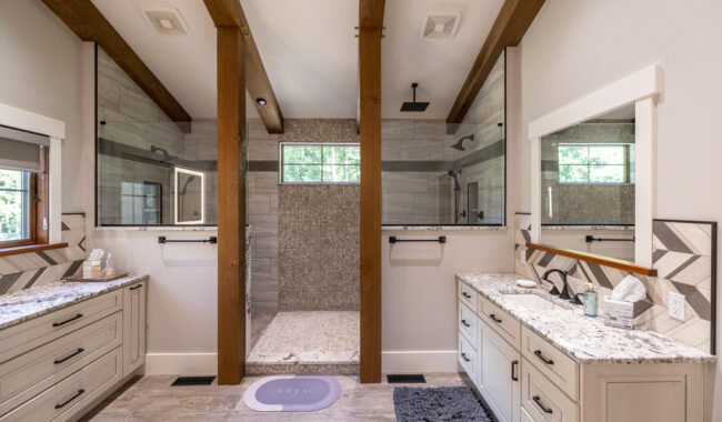 Modern bathroom with a large, open walk-in shower in the center, flanked by two separate vanities with granite countertops on each side. Exposed wooden beams, neutral tones, and natural light from windows create a spacious feel.